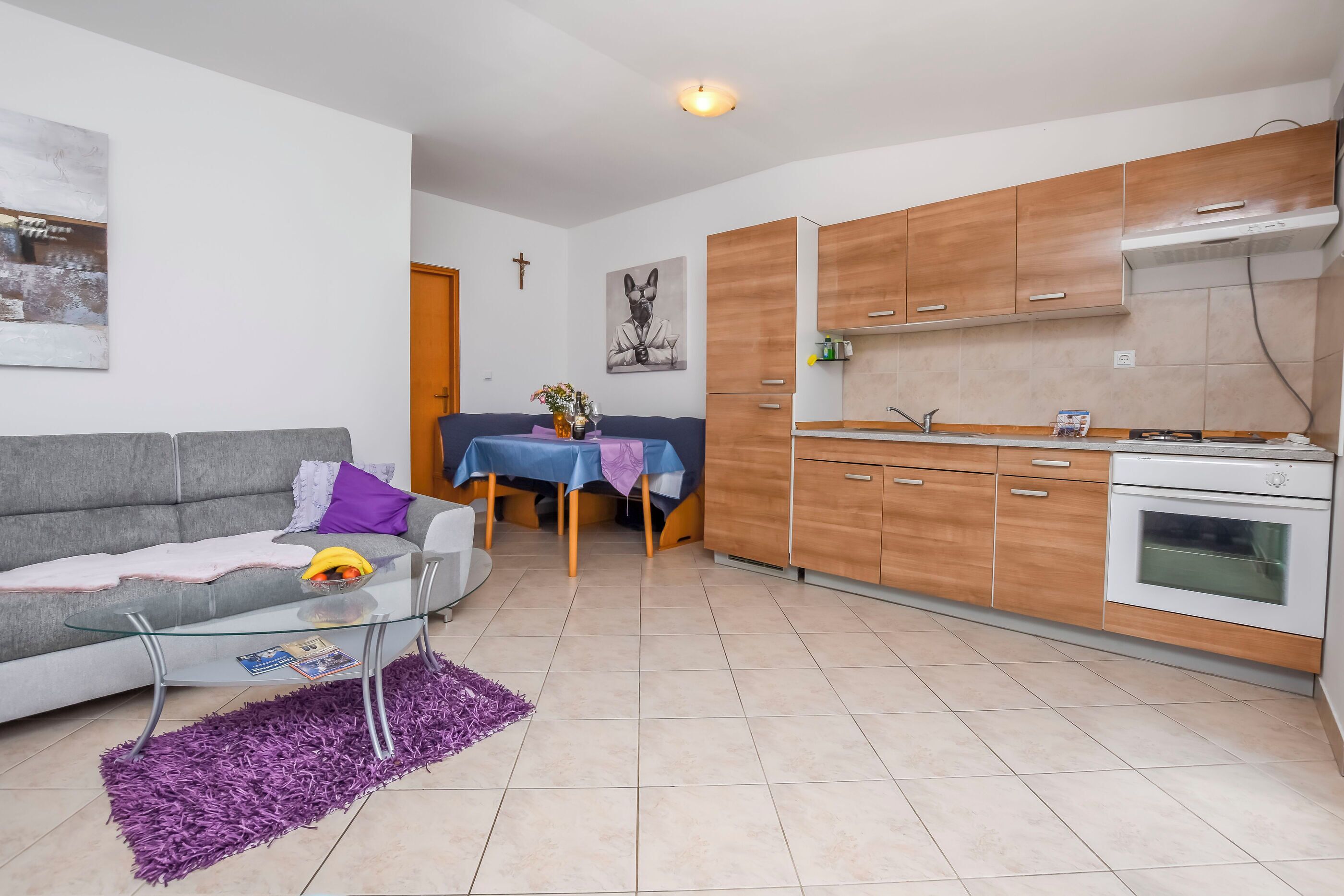 Un salon moderne avec un canapé gris, une table basse en verre et un tapis violet. La cuisine est équipée de meubles en bois et d’un four blanc.