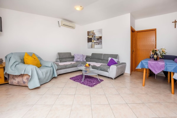 A living room with a sectional sofa, a glass coffee table, and a dining table with a purple tablecloth. - Apartments Kata (Rogoznica)