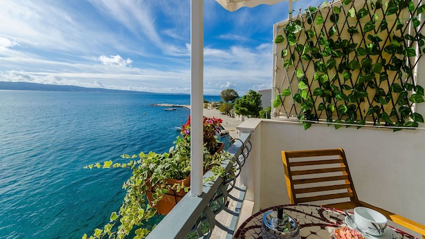 Un balcon avec vue sur la mer, une chaise en bois, une table avec un bol et une soucoupe, et des plantes en pot.