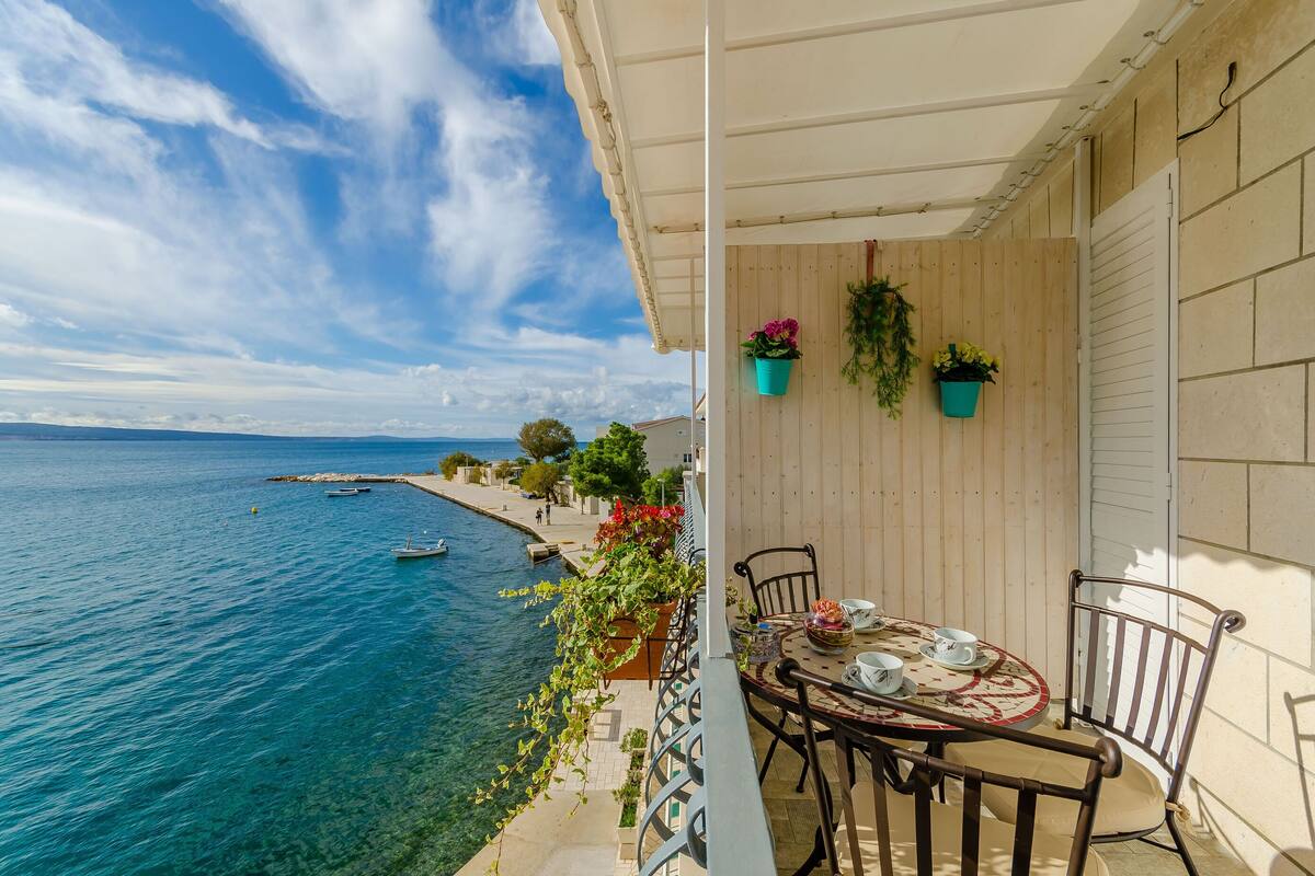 Ein Balkon mit Tisch und Stühlen mit Blick auf eine Küstenlandschaft, blaues Wasser und Boote.