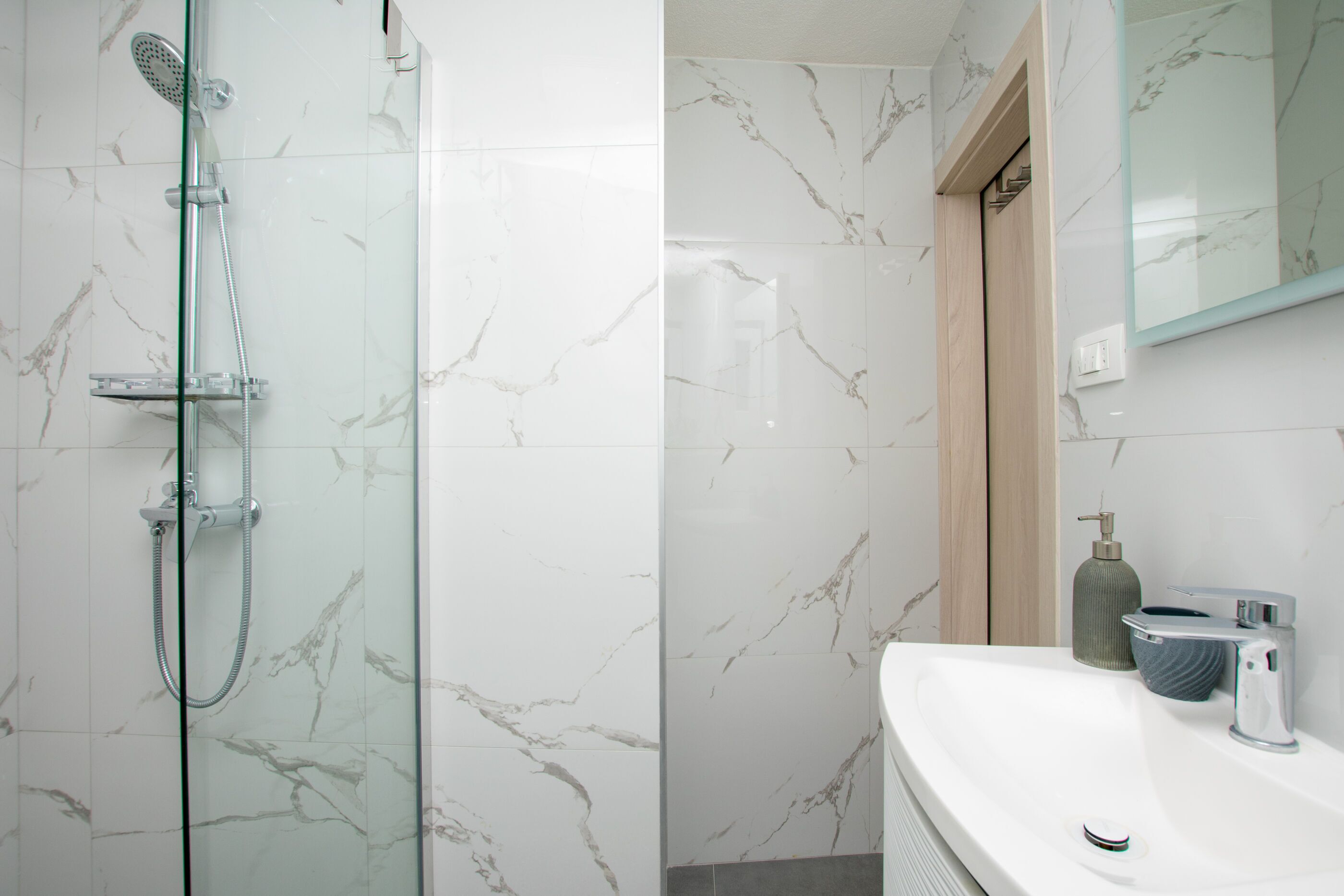 A modern bathroom with a glass-enclosed shower, marble walls, a white sink, and a mirror.