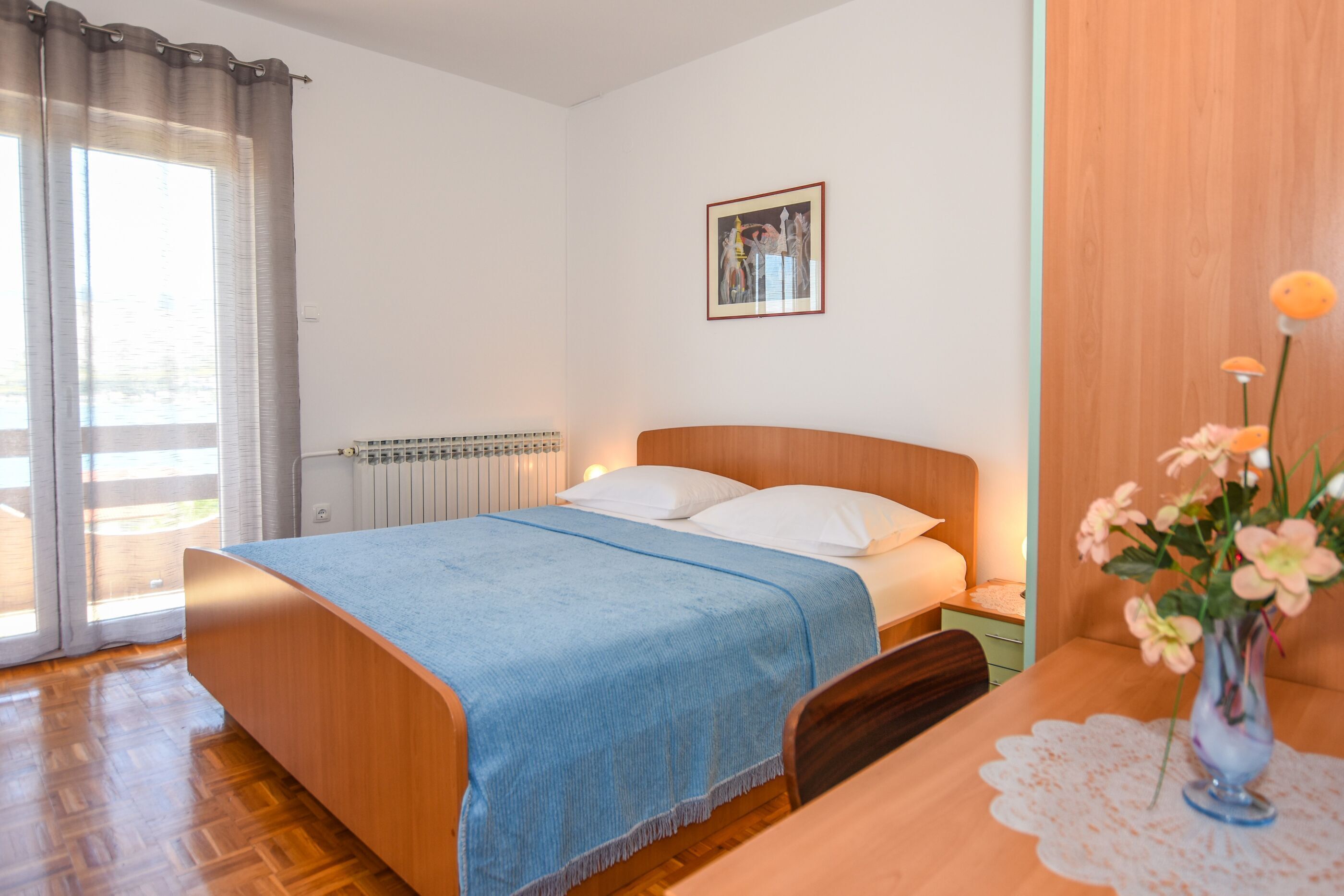 Une chambre à coucher avec un lit en bois, une couverture bleue, une table de chevet ornée d’un vase de fleurs et un radiateur.