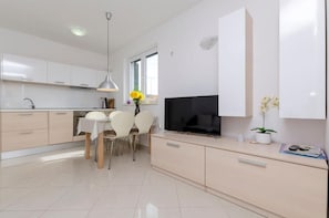 A modern kitchen with white cabinets, a dining table with chairs, a TV, and a window with flowers. - Apartments Dragica (Vodice)