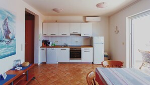 A compact kitchen with white cabinets, a dishwasher, an oven, and a refrigerator. There is a dining table with chairs, a bench, and a painting on the wall. - Apartments Jure (Okrug Gornji)