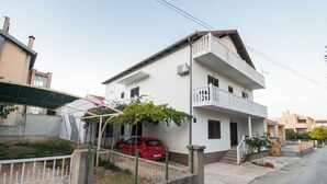 A two-story residential building with a balcony, a red car parked in front, and a small garden area. - Apartment Mijo (Vodice)