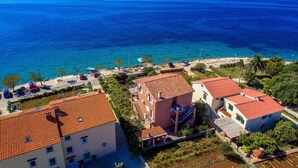 A coastal building with a red-tiled roof, adjacent to a beach with clear blue waters and a distant mountain. - Apartments Ivan Sea View Lun (Lun)