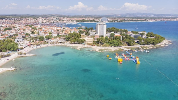 Luftaufnahme einer Küstenstadt mit klarem türkisblauem Wasser, einem Strand und einem großen Gebäude im modernen Stil.