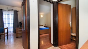 Ein Zimmer mit einem Bett, einem Schreibtisch und einem Stuhl. Eine Holztür und ein Vorhang sind ebenfalls vorhanden.