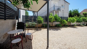 Ein überdachter Außenbereich mit Tischen und Stühlen, ein Gebäude mit Balkon und ein Kiesgrundstück.