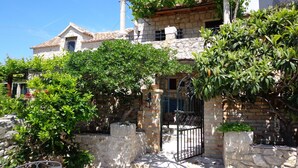Un edificio in pietra con tetto in tegole, un cancello in metallo e rigogliosa vegetazione.