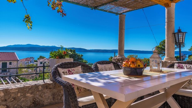 Una terrazza con mobili in vimini, un tavolo con della frutta e vista mare e montagne.