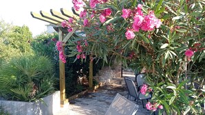 Un patio in pietra con una pergola fiorita di fiori rosa, arredi da esterno e un'area giardino con diverse piante.