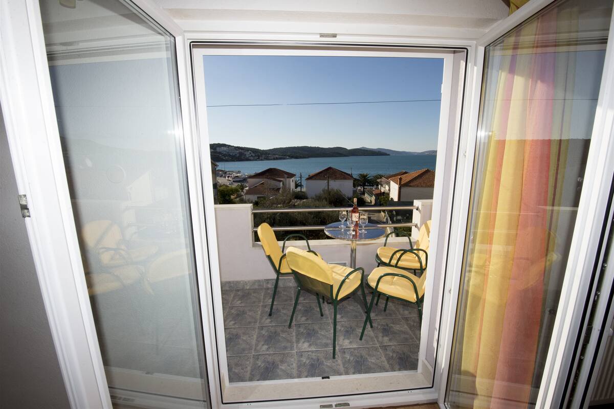 A balcony with a table and chairs, overlooking a coastal area with houses and a clear sky.