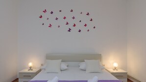 Un lit bien fait, recouvert d’une couvre-lit violette, avec deux oreillers blancs et deux tables de chevet blanches, chacune surmontée d’une lampe.