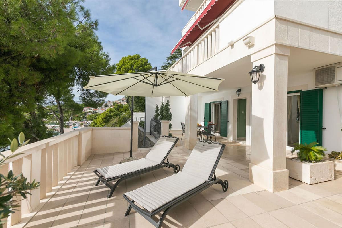 A balcony with two lounge chairs and a large umbrella, a building with green shutters, and a view of the surrounding area.