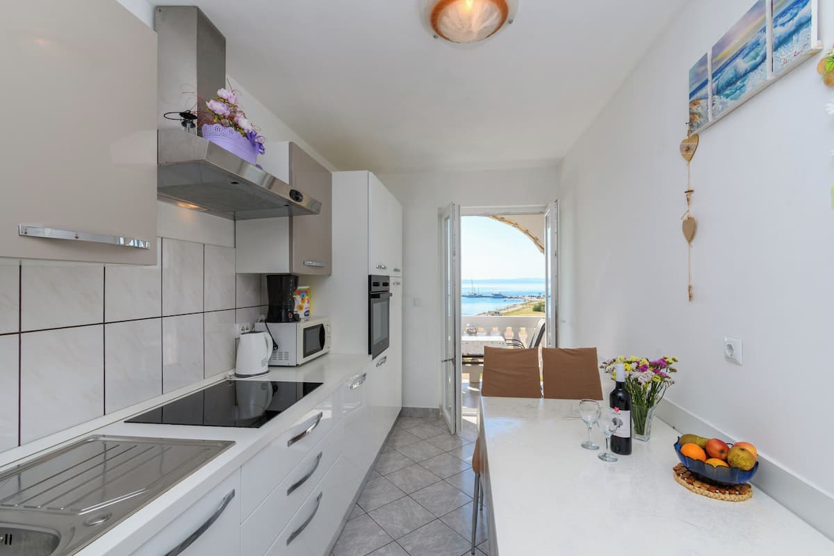 A modern kitchen with a view of the sea, a dining area with a table set for two, and a small fruit bowl on the counter.