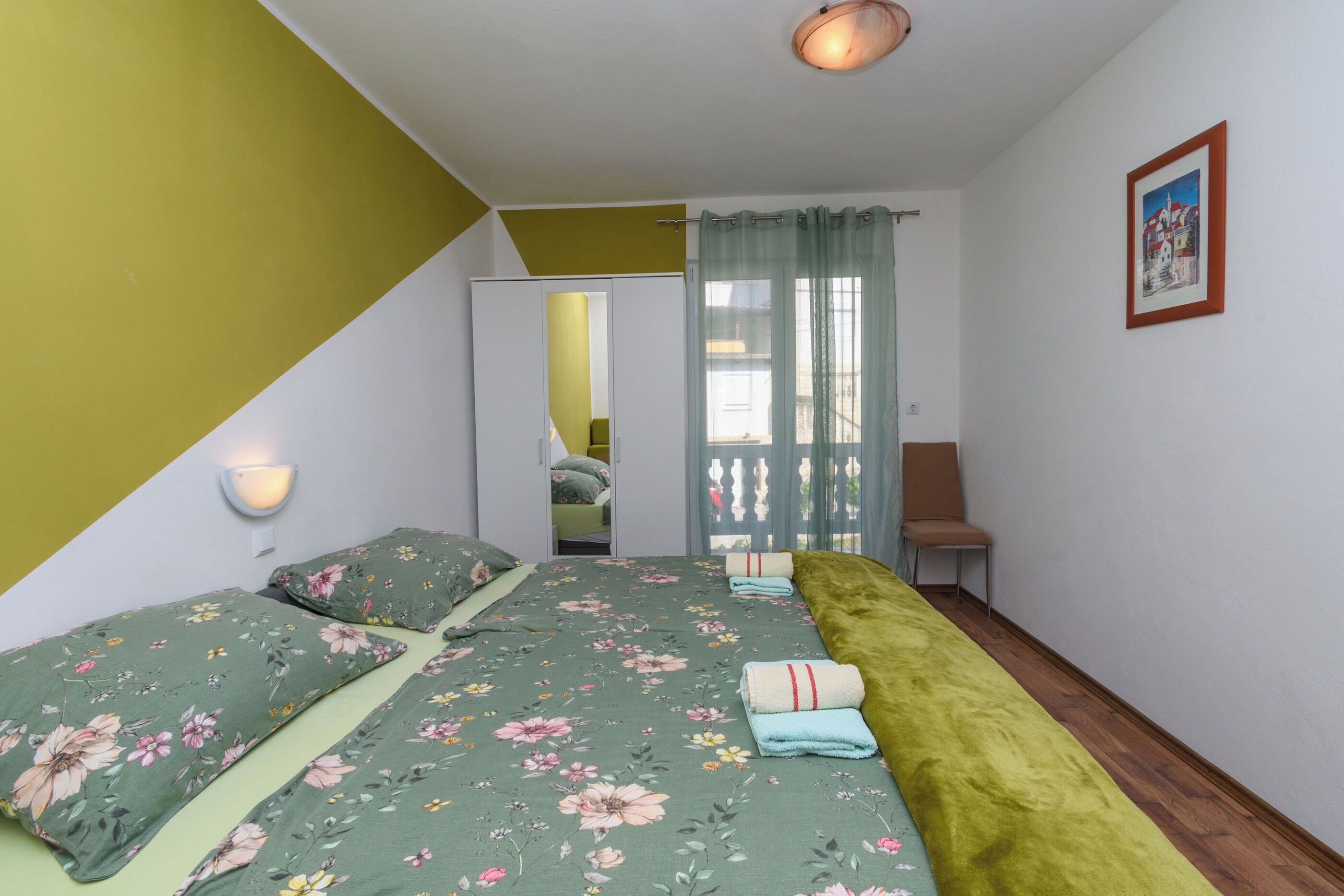 Ein Schlafzimmer mit Bett, Sessel, Kleiderschrank und einem Fenster mit Vorhängen.