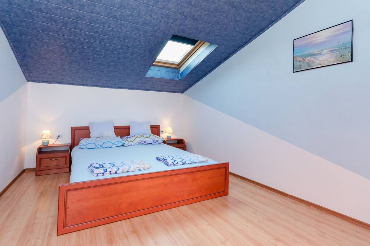 A bedroom with a skylight, a bed with a patterned bedspread, bedside tables with lamps, and a framed picture on the wall.