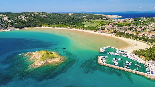 An aerial view of a coastal area with a marina, sandy beach, and residential buildings. - Apartments Albina (Lopar)