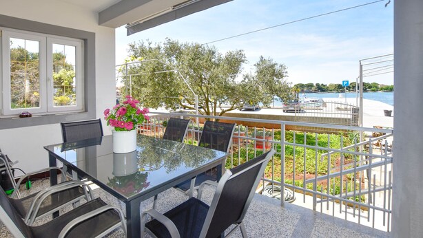 A balcony with a glass table and chairs, a vase of flowers, and a view of the sea and trees. - Apartment Danica (Sukosan)