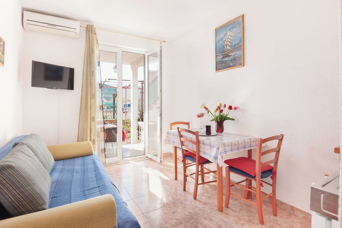 A living room with a sofa, a dining table with chairs, a TV mounted on the wall, and a painting.