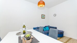 Un coin repas avec une table blanche, un plateau sur lequel se trouvent une bouteille et des verres, une petite plante et un canapé bleu à rayures agrémenté de coussins.