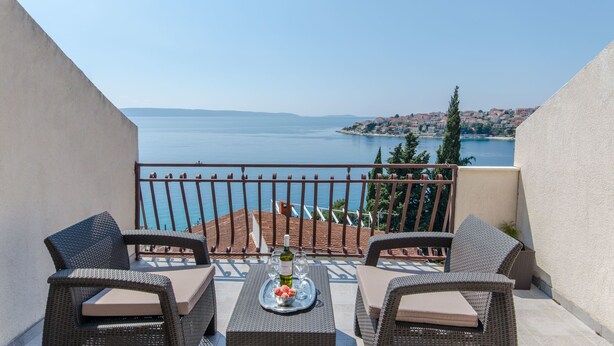 Un balcon agrémenté de meubles en osier, un plateau avec de la nourriture et des boissons, et une vue sur la mer et les bâtiments éloignés.