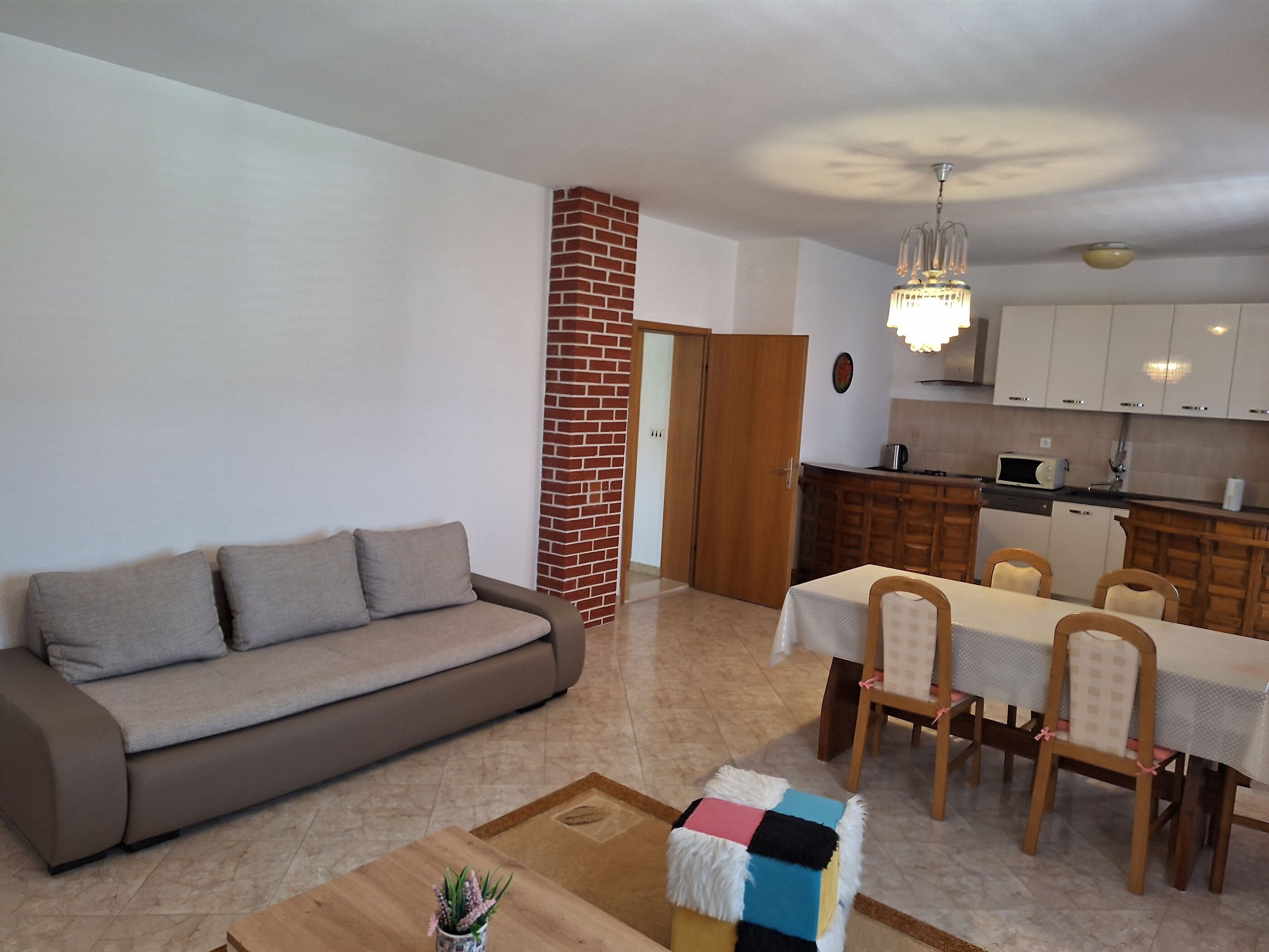 A living room with a sofa, a dining table with chairs, a kitchen area, and a chandelier.