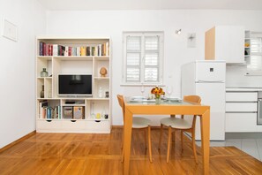 A modern living space with a wooden floor, a dining table with chairs, a bookshelf with a flat-screen TV, a window with blinds, and a kitchen area with white cabinets. - House Urban Bliss (Vodice)