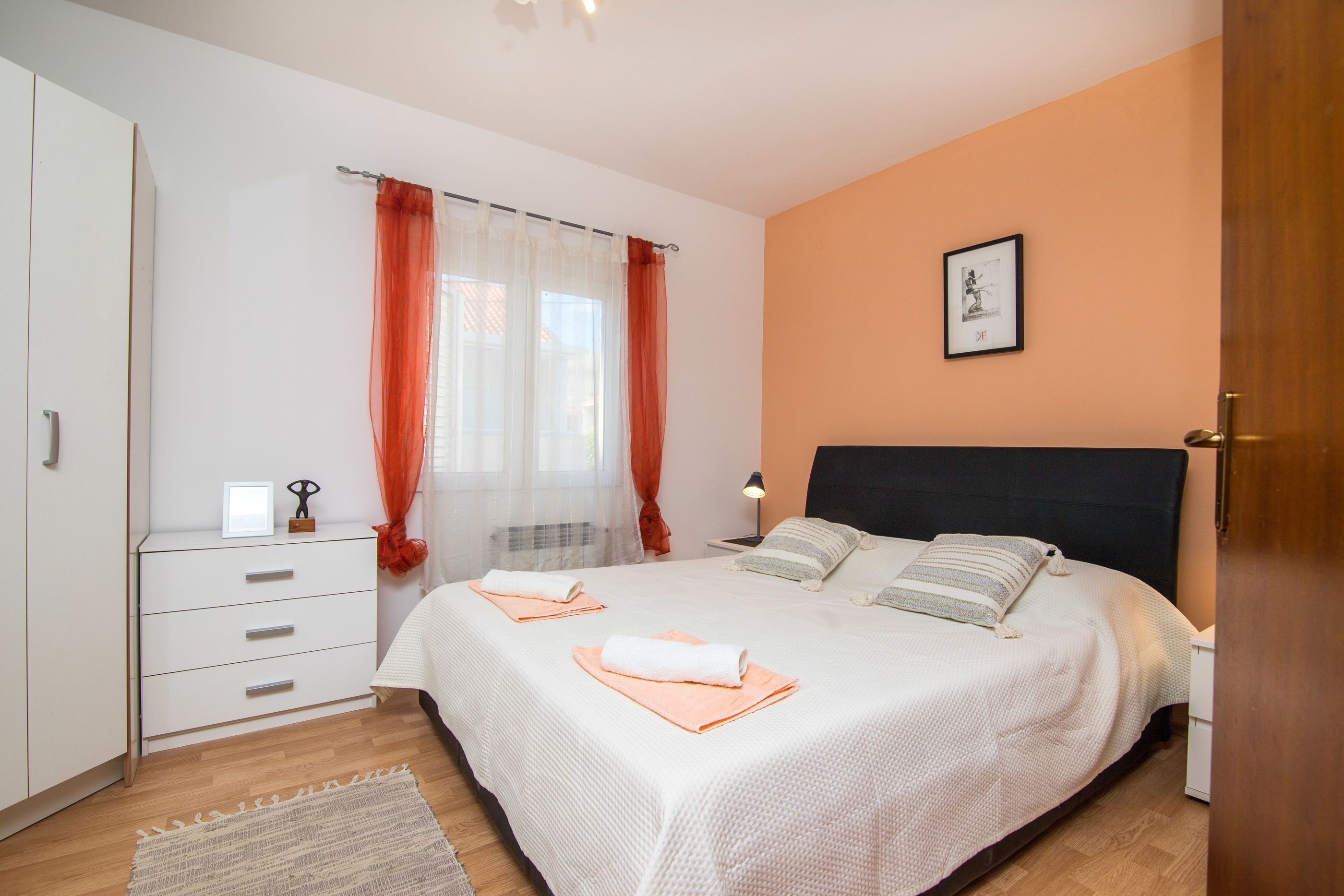 A bedroom with a bed, a nightstand, a dresser, a window with curtains, and a framed picture on the wall.
