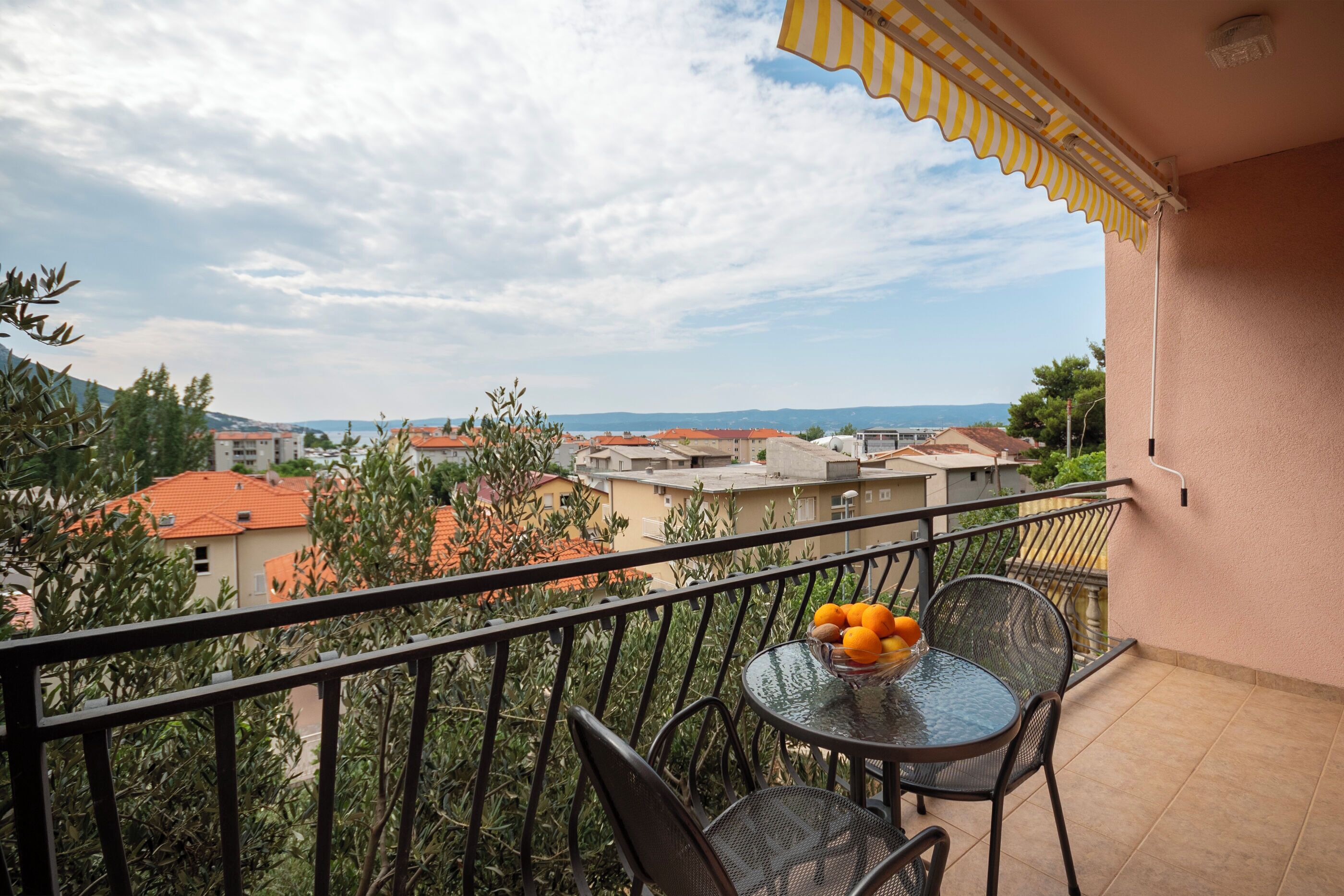 A balcony with a table and chairs, a yellow-striped awning, and a view of buildings and trees.