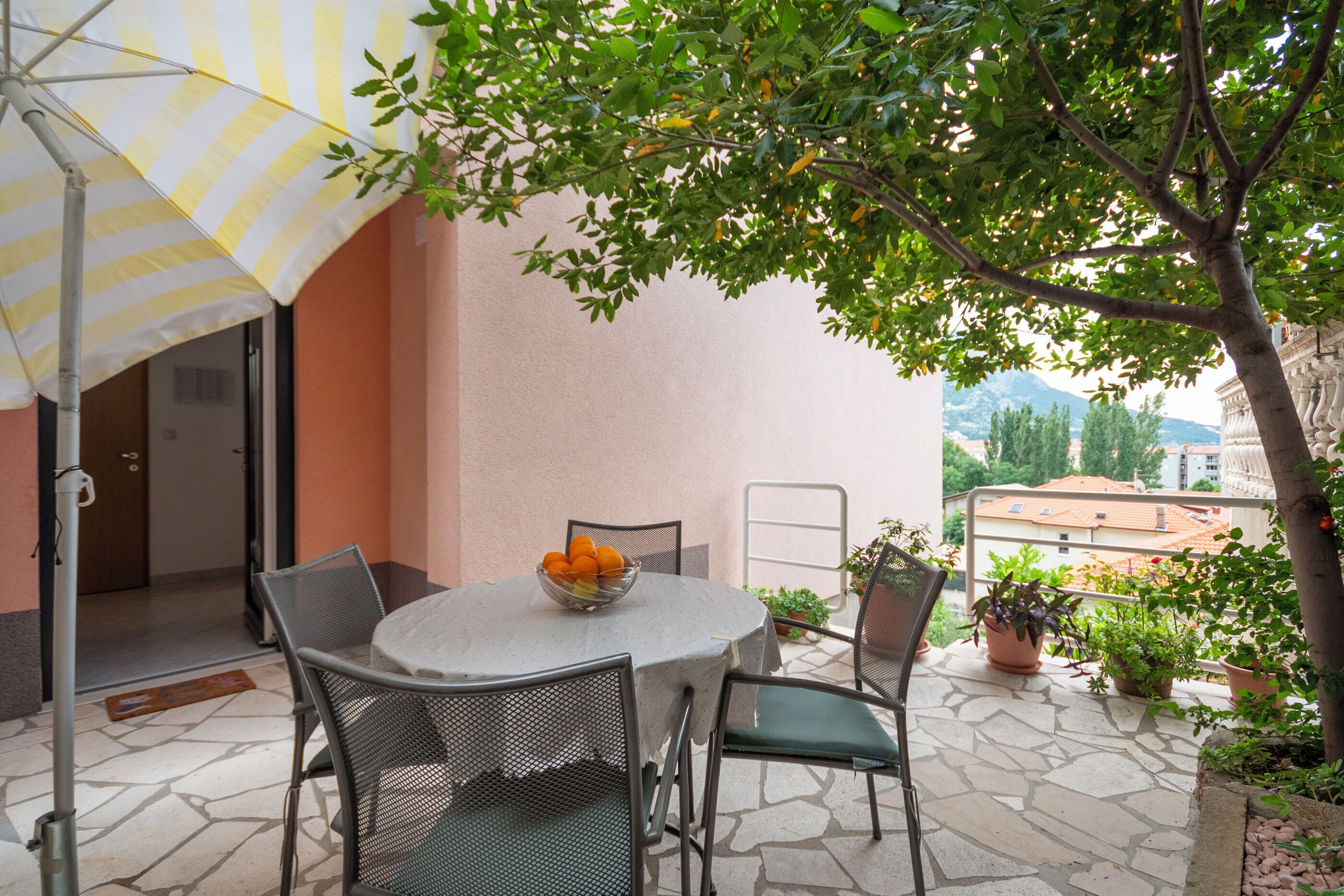 A round table with chairs and a bowl of fruit, an umbrella, and potted plants on a balcony with a view of buildings.