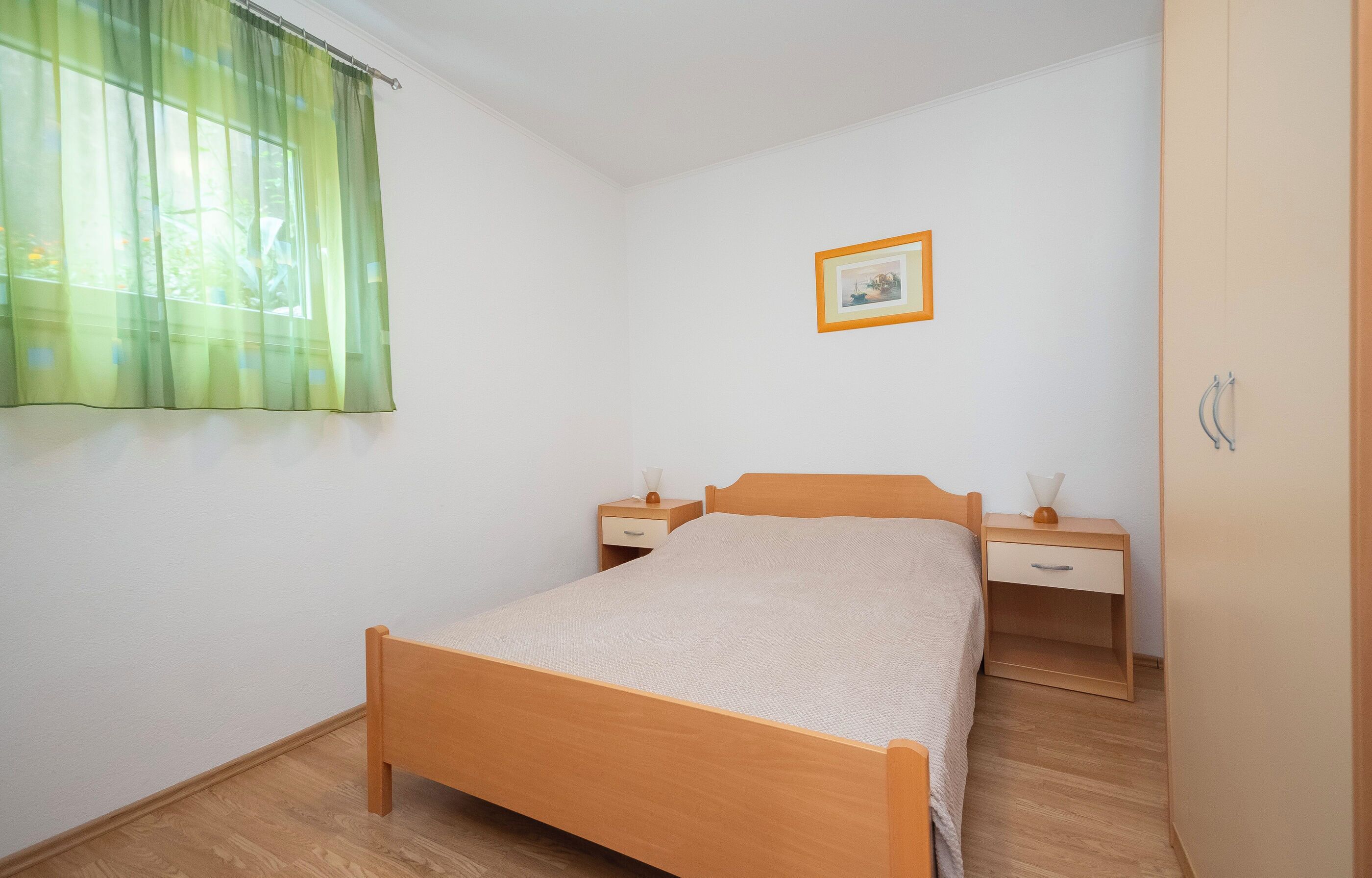 A bedroom with a wooden bed, two bedside tables, a nightstand, a framed picture on the wall, and a window with sheer curtains.