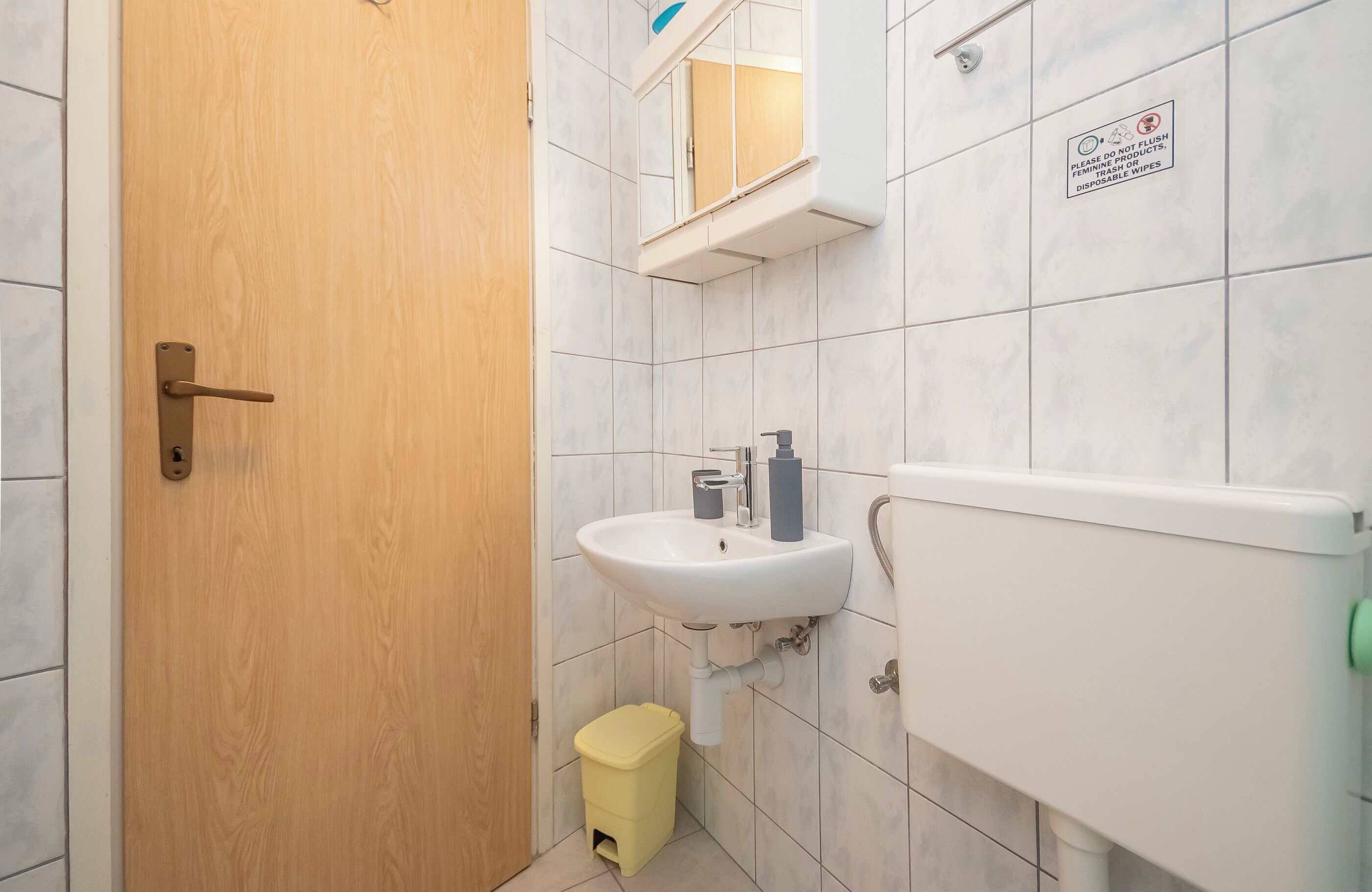 Une salle de bain avec une porte en bois, un lavabo blanc avec un miroir au-dessus, et des toilettes. Il y a une poubelle sous le lavabo.