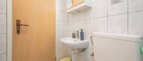 Une salle de bain avec une porte en bois, un lavabo blanc avec un miroir au-dessus, et des toilettes. Il y a une poubelle sous le lavabo.