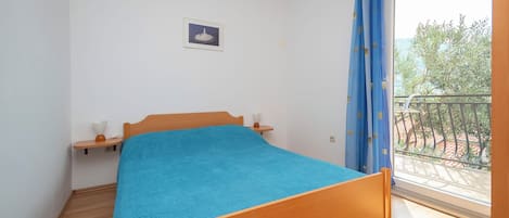 Een slaapkamer met een houten bed, een klein nachtkastje, een aan de muur hangend schilderij en een balkon met uitzicht op bomen en een hekwerk.
