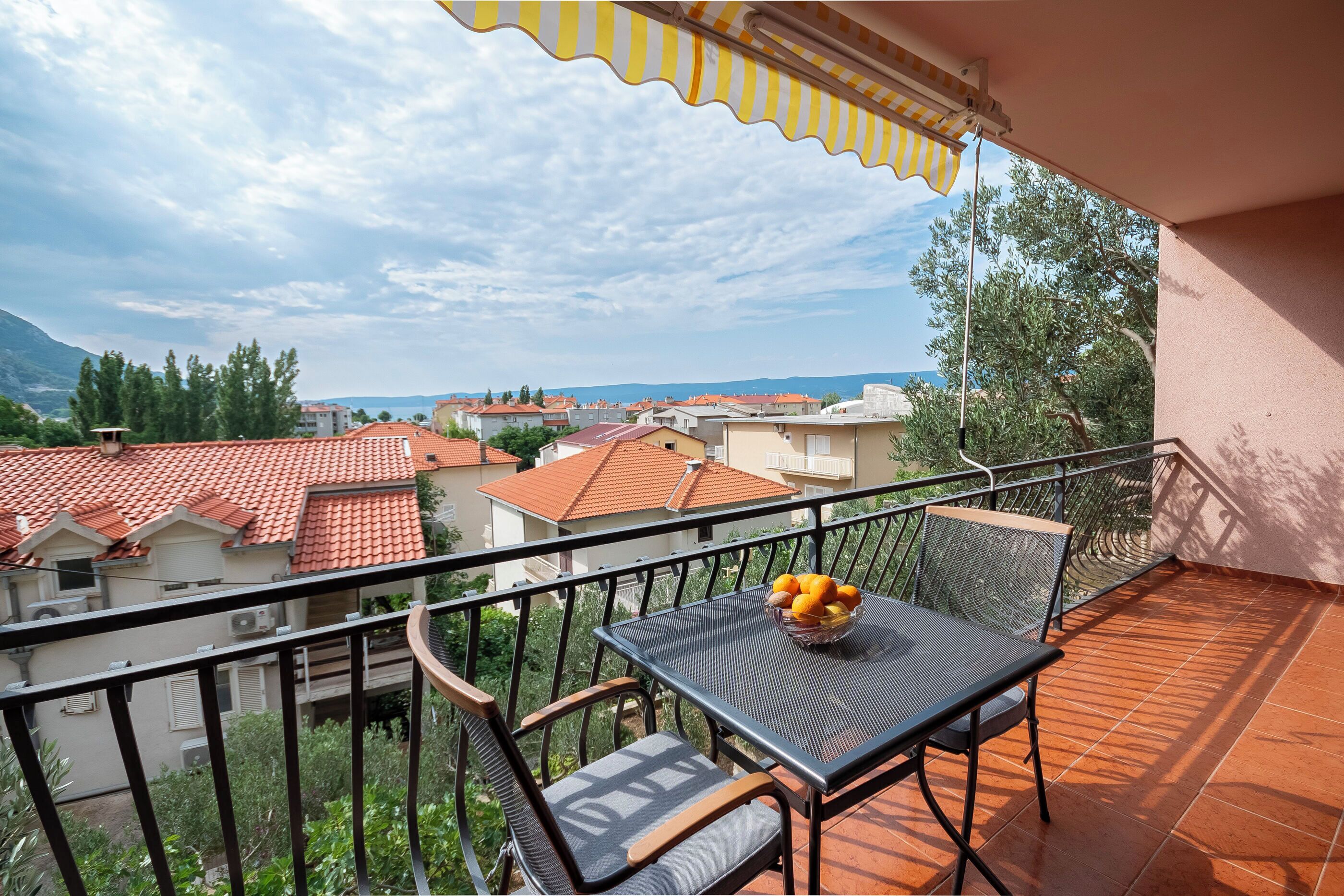 A balcony with a table and chairs, a basket of fruit, and a view of a town with red-roofed buildings and greenery.