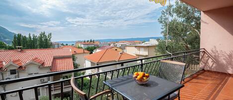 A balcony with a table and chairs, a basket of fruit, and a view of a town with red-roofed buildings and greenery.