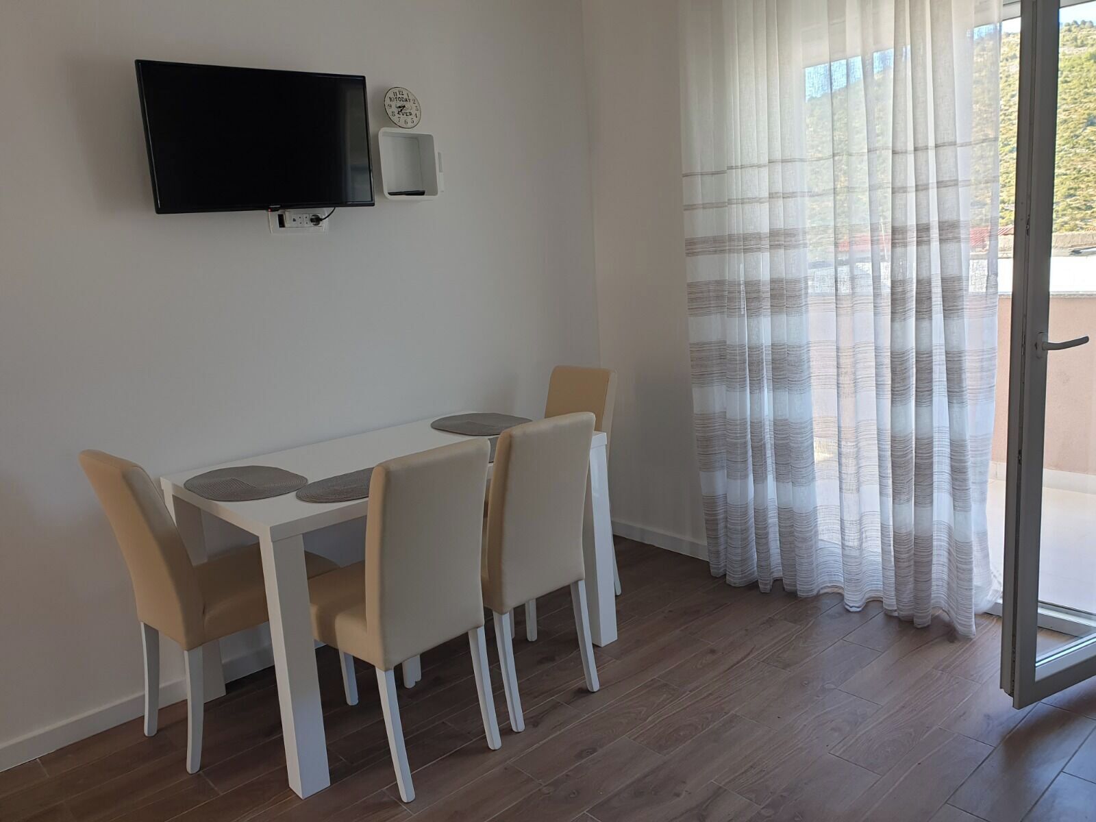 Ein Essbereich mit weißem Tisch und Stühlen, einem flachbildfernseher an der Wand, einer kleinen Uhr und Blick durch ein Fenster mit durchsichtigen Vorhängen.