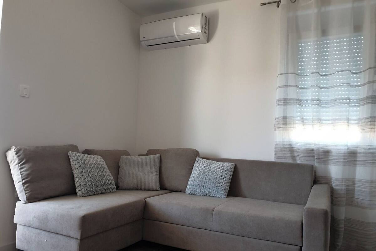 A beige sectional sofa with patterned cushions, a wall-mounted air conditioner, and a window with striped curtains.