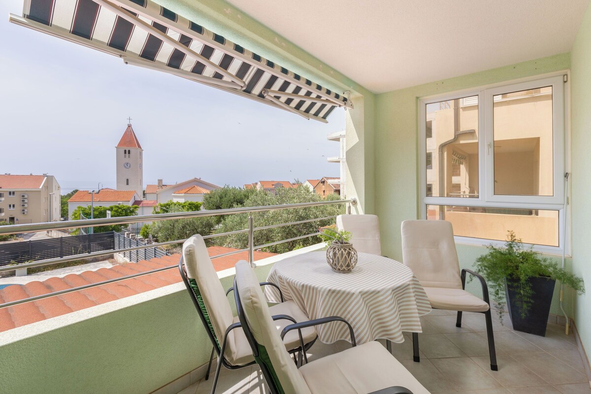 A balcony with a table and chairs, a potted plant, and a view of buildings and a tower.