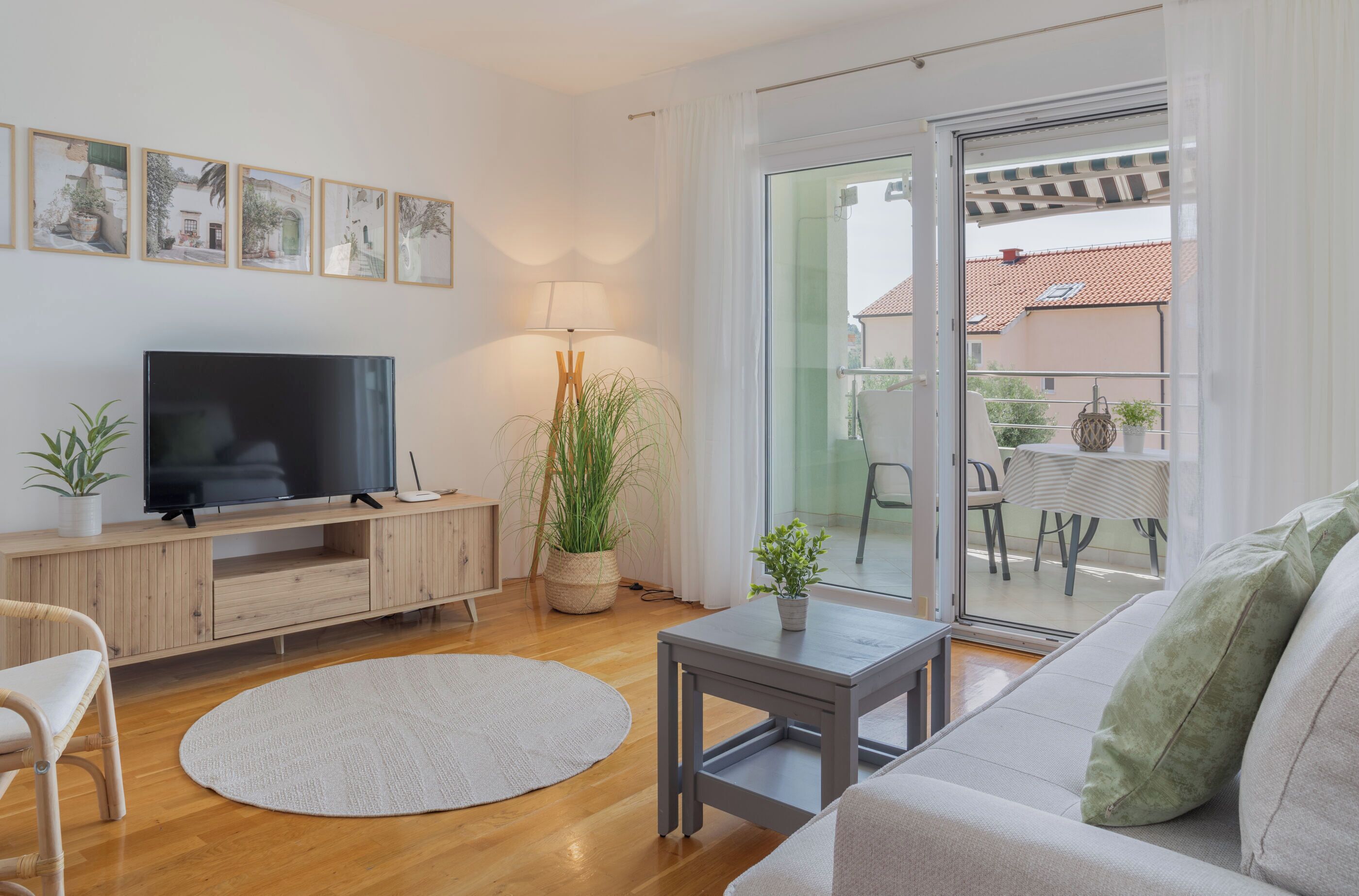 A modern living room with a flat-screen TV, wooden furniture, a round rug, and a balcony with outdoor seating.