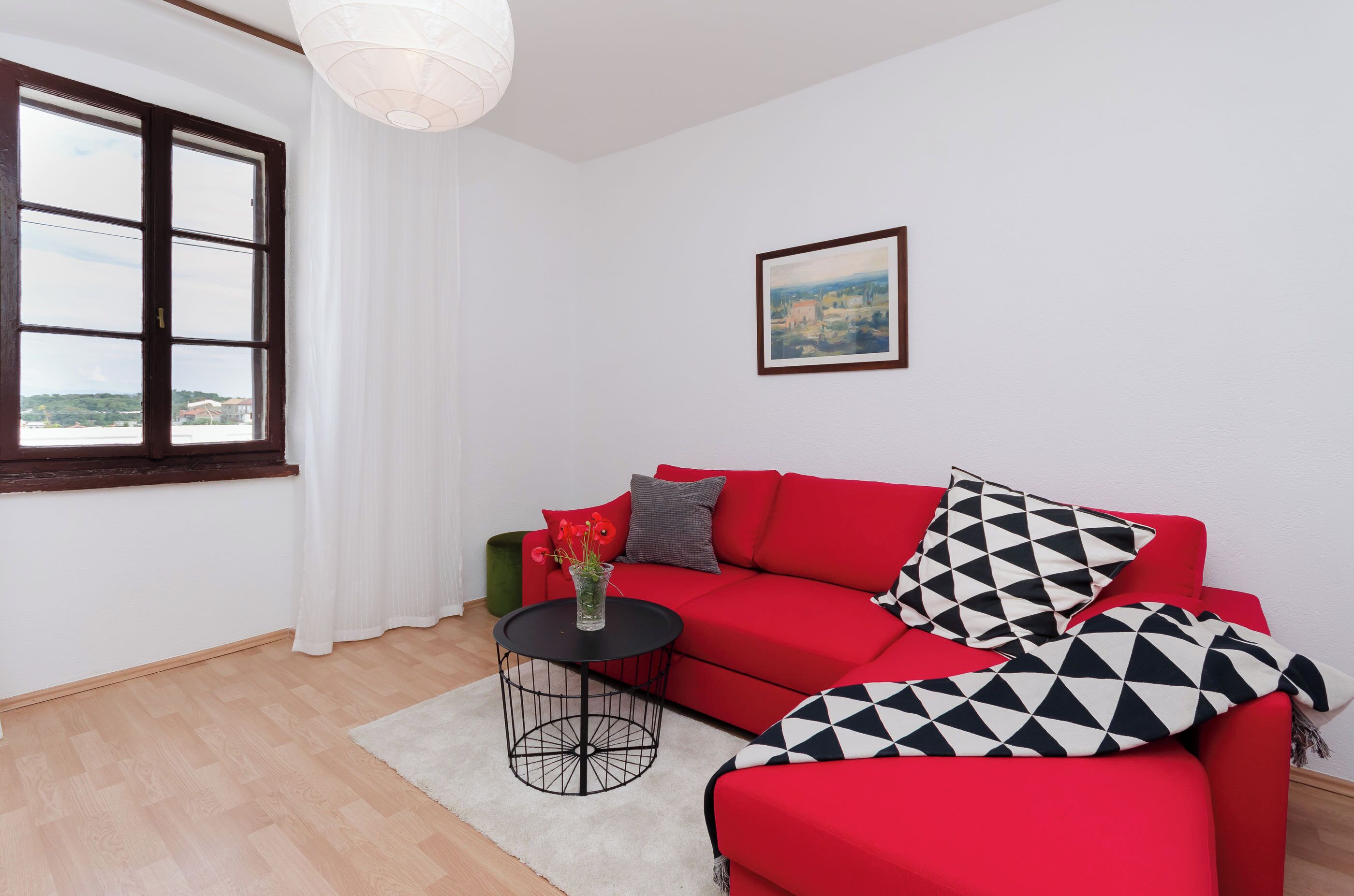 A red sofa with geometric pillows, a small round table with a vase, and a framed picture on the wall.