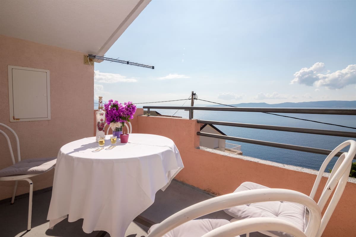 Un balcon avec une table dressée pour deux personnes, offrant une vue sur une étendue d’eau et des collines lointaines.