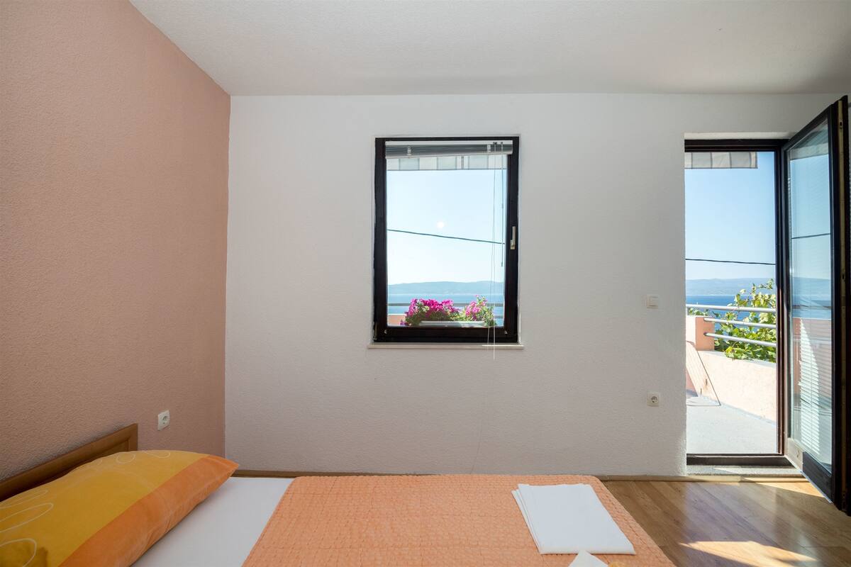 Une chambre avec un lit, une fenêtre donnant sur la mer et une porte ouverte sur un balcon.