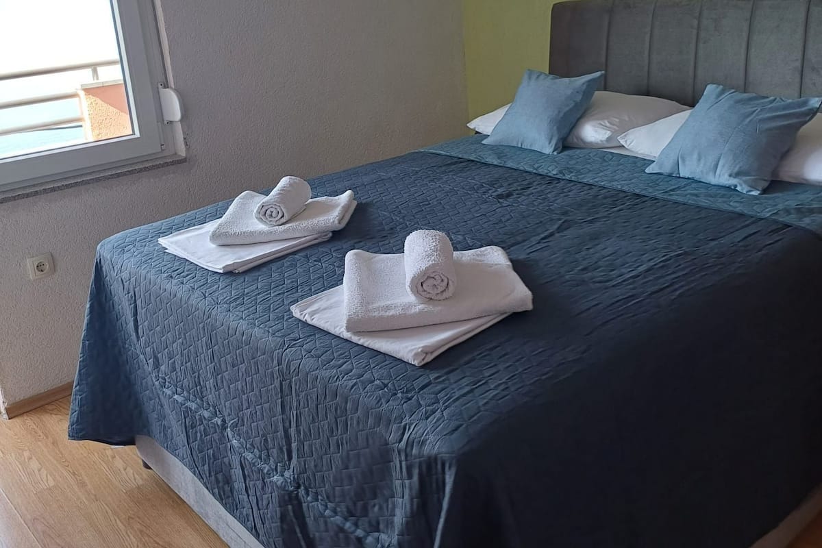 Un lit bien fait, avec une couvre-lit bleue et des oreillers blancs, une fenêtre avec des stores et un plancher en bois.
