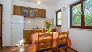 A kitchen with wooden cabinets, a white refrigerator, a dining table with a checkered tablecloth, and a window with a view of trees. - Apartments Gordana (Mimice)