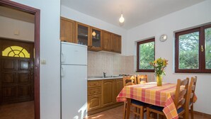 A kitchen with wooden cabinets, a white refrigerator, a tiled backsplash, a dining table with chairs, and a window with a view of trees. - Apartments Gordana (Mimice)
