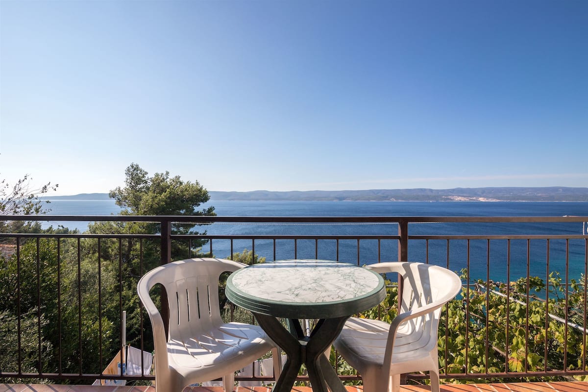 A balcony with a table and two chairs overlooking a body of water and distant hills.