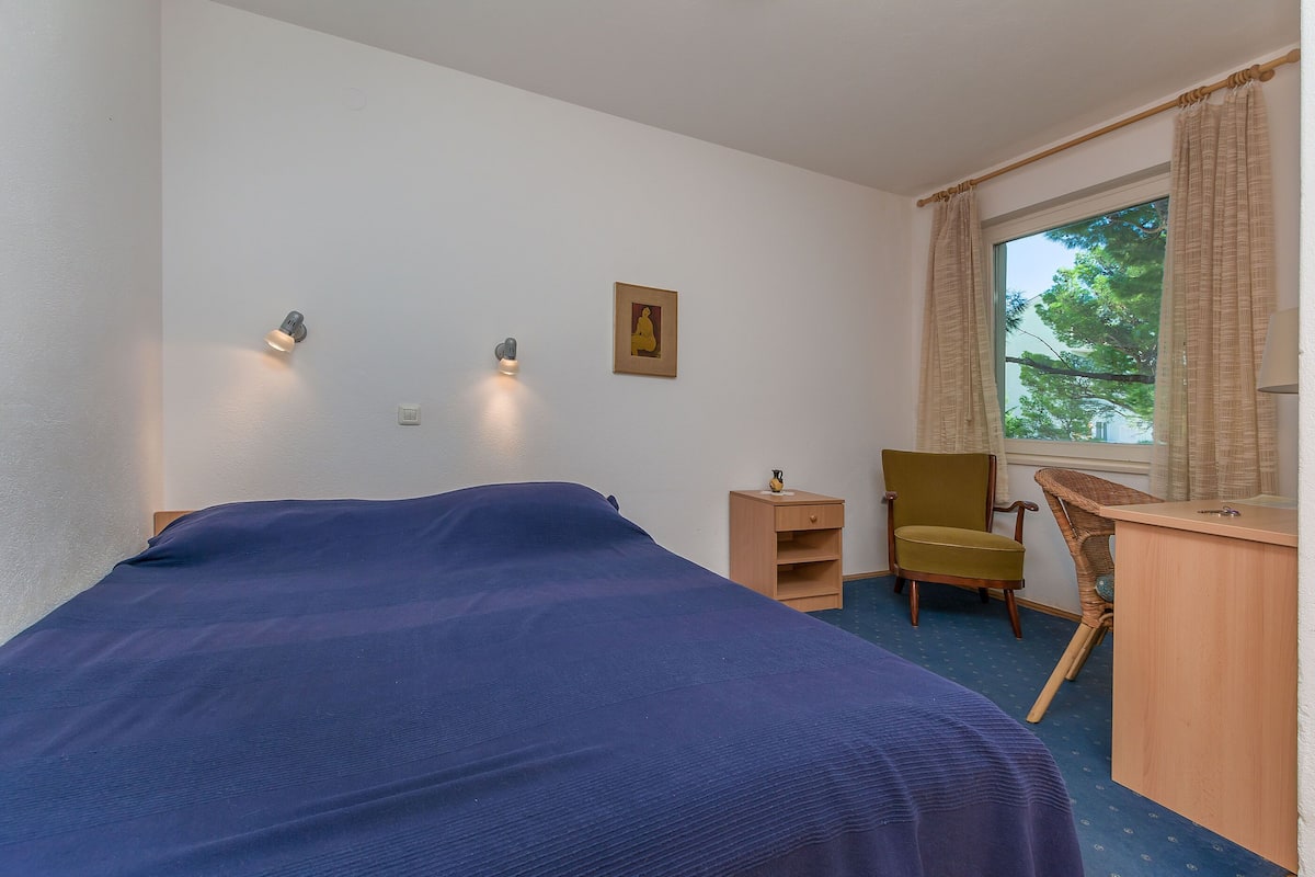 A bedroom with a bed, a desk, a chair, a small cabinet, and a window with curtains.